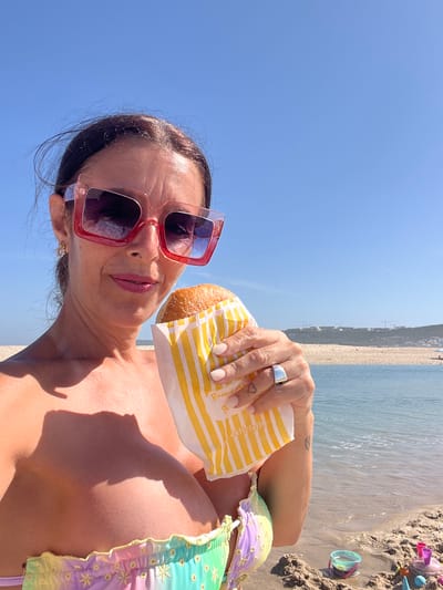 The image shows a woman on a beach, holding a pastry in a paper bag. She is wearing large, pink-framed sunglasses and a colorful bikini top. The background features a blue sky, a body of water, a sandy beach, and a distant coastline with buildings. It is daytime and the sun is casting shadows. The setting appears to be Foz do Arelho beach, located in the municipality of Caldas da Rainha, Portugal, known for its lagoon that meets the Atlantic Ocean.