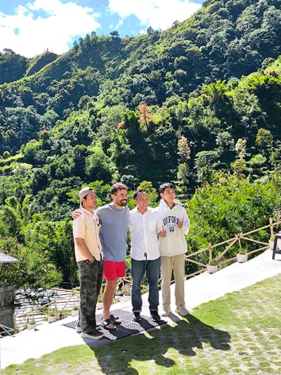 Four men stand together outdoors. The man on the left wears a tan button-down shirt, camouflage pants, and a cap. Next to him, a man with a beard wears a gray shirt and red shorts. The third man wears a white shirt and jeans. The man on the right wears a gray hoodie with the word "OXFORD" on it and tan pants. All men are smiling. The two men in the middle have their arms around each other. The man on the right is giving a thumbs up. They are standing on a stone walkway. In the background, there is a lush green mountain covered with trees. The sky is blue with some white clouds. They are in Sembalun Bumbung, Indonesia. A wooden fence and some potted plants are visible near the edge of the walkway. A small structure is seen behind the man on the left.
