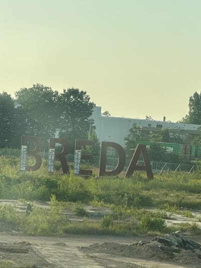 This media file captures a weathered, large metal sculpture spelling out "BREDA" in a vertical orientation. The letters are rusty and show signs of wear and tear, with some parts appearing to be filled with a lighter colored material. The sculpture stands outdoors in a grassy, somewhat overgrown area, with a dirt path visible to the right. In the background, a large, modern building with a light-colored facade and some green shipping containers are visible. Trees and lush vegetation surround the building and the sculpture. A temporary fence is also present behind the sculpture. The lighting suggests it might be late afternoon or early morning, with a soft, slightly hazy sky. The overall impression is one of an industrial or post-industrial landscape, with the sculpture adding a striking artistic element. The text on the sculpture reads "BREDA". The location context indicates this scene is in Wuustwezel, Belgium.