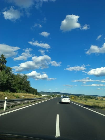 The image shows a view from the driver's seat of a vehicle traveling on a highway. The highway has a black asphalt surface with clear white lane markings. To the left of the highway, there is a guardrail and a grassy embankment with trees and vegetation. In the distance, there are hills and a small town or village visible. The sky is a clear blue with scattered white clouds. A white car is visible ahead in the right lane of the highway. A yellow sign with black text is also visible on the right side of the highway. The image is taken during daylight hours. The location is Krašovice, Czechia.