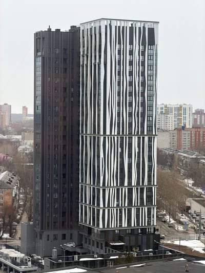 An elevated, distant perspective captures an urban scene in Gamovo, Russia, under an overcast sky, indicating a cool or cold climate with bare trees and visible snow patches on the ground. The foreground is dominated by two modern high-rise buildings. The structure on the left is a tall, slender building with a dark brown or bronze facade, featuring numerous rectangular windows and vertical paneling, topped with a flat roof that includes minor protrusions. Directly adjacent to it on the right is a multi-story building characterized by a dark facade interlaced with prominent, wavy, vertical white architectural elements that segment its exterior. This building also has numerous rectangular windows.
At the base of these main structures, a lower-level grey building complex with flat roofs and several visible outdoor air conditioning units is present. Patches of white snow are scattered on the ground and lower rooftops. To the right, a multi-lane asphalt road runs diagonally, with several vehicles including cars and a red bus visible in traffic. Bare deciduous trees line this road. A narrower, less trafficked street is visible to the left, with at least one parked white car. A few human figures are discernible walking on the sidewalks. The background reveals a dense urban landscape comprising numerous residential and commercial buildings of varying heights and architectural styles, extending towards the horizon under the uniform grey sky.