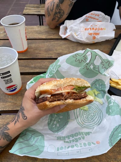 A person with tattoos is holding a burger on a wrapper with green tomato shapes in their right hand. A cup of Pepsi is next to the burger, as is another empty cup. There is a wrapper for Jollo Jr. chicken, and some fries, on the table, which is made of horizontal wooden slats. The location is identified as Monte Grande, Argentina.