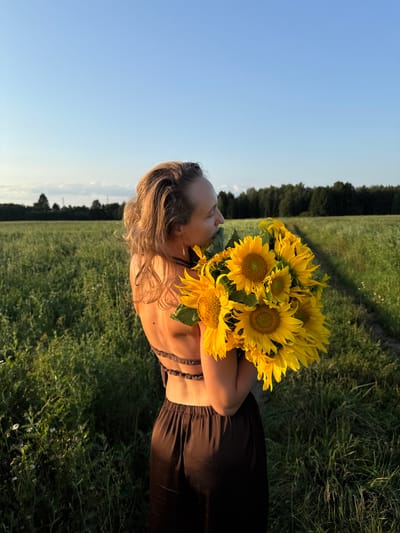 In Aksaitovo, Russia, a woman stands in a field of green grass, holding a bouquet of sunflowers. She is wearing a dark brown dress with an open back. The sun is setting, casting a warm glow on the scene. The sky is clear and blue, with a few clouds in the distance. The field is bordered by a forest.