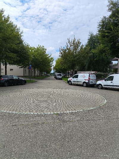 The image presents a static street scene in Wolfisheim, France, under a sky with scattered white clouds. The foreground consists of dark asphalt pavement, leading to a central circular paved area composed of light grey interlocking pavers arranged in concentric rings, outlined by a white line. A tree-lined street extends into the background, flanked by buildings. On the left side of the street, a black SUV is parked near a grass verge. A blue circular road sign with a red diagonal line is visible on a pole near the SUV. On the right side, three white commercial vans are parked along the street. The closest van displays "nex." branding on its side and features a roof rack with red and white safety stripe markings. Additional vehicles are visible in the distance along the street. White multi-story buildings are present on the left, and a multi-level structure is visible on the right, further down the street. No individuals are present.