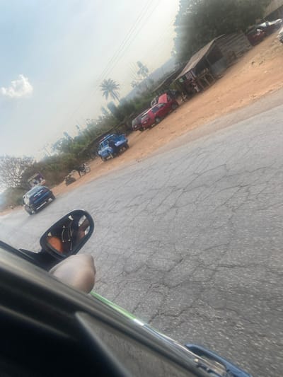 The image displays an oblique aerial perspective of a roadside scene. Several vehicles are parked along a dirt shoulder next to a cracked asphalt road. Vegetation is visible to the left, including a palm tree. A partial view of a vehicle's interior, including the side mirror and what appears to be the driver's arm, is visible in the lower right corner. The photograph appears to have been taken from inside a moving vehicle. The overall image quality is somewhat blurry, possibly due to motion.