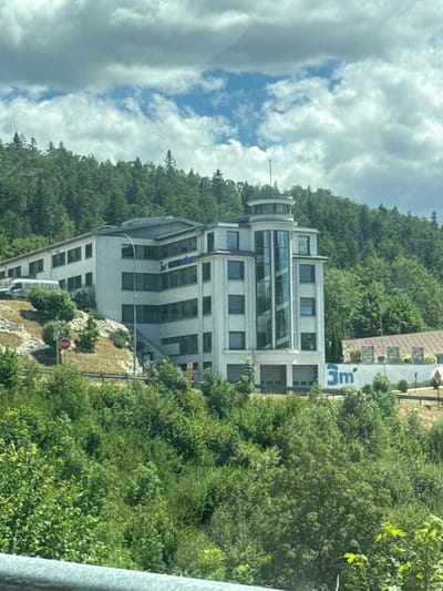 The image shows a modern, multi-story white building with a distinctive circular glass tower on the right side, situated on a hillside covered with lush green trees. The building features numerous windows and appears to be an office or commercial space. A "STOP" sign is visible on the left side of the image, near a road. In the background, more trees and a cloudy sky are present. The building has a sign with blue lettering that reads "comestibles" on the left side and a prominent blue logo with "3m" on the right side. The surrounding environment suggests a semi-rural or suburban setting.