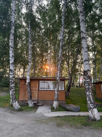 This is a photograph of a small wooden cabin with a numbered "15" on its exterior, situated amongst a grove of birch trees. The cabin has a wooden door, a window with white curtains, and a small wooden deck leading to its entrance. The sun is setting, casting a warm glow through the trees and onto the cabin. An inflatable boat lies on the deck next to the cabin. The scene appears to be in a natural setting, possibly a campsite or a rural area, consistent with locations found in Parnaya, Russia, known for its natural beauty and recreational areas. The overall atmosphere is serene and peaceful. There are no people visible in the immediate foreground or interacting with the cabin.