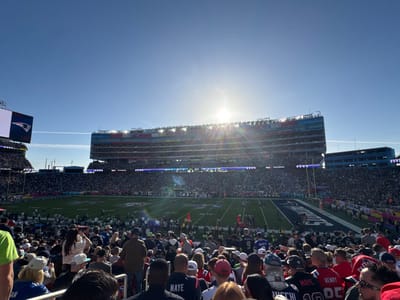 A sun-drenched stadium packed with fans, likely for a sporting event, possibly football, is shown. A large screen displays a team logo. The crowd is dense, a mix of people wearing casual clothes and jerseys. The field is visible between the spectators. The sun is high in the sky, creating glare over the top tier of seats.