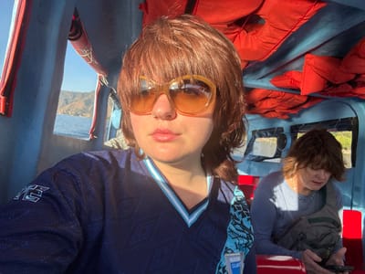 The image displays two individuals inside a passenger boat on Lake Atitlán, near San Pablo La Laguna, Guatemala. The boat's interior features blue walls and ceiling, with red seating. Several folded red life jackets are stored in overhead compartments.

In the foreground, a female with reddish-brown, shoulder-length hair and bangs wears large, rounded amber-tinted sunglasses. She has a small ring in her left nostril. She is wearing a dark blue long-sleeved shirt with a light blue V-neck trim and a subtle geometric pattern. A partial "SELECCIÓN GUATEMALA" logo is visible on the lower right of her chest, along with a light blue and black patterned cloth draped over her left shoulder. She is looking directly towards the camera with a neutral expression.

In the background, to the right, a second female with short, dark brown hair is seated on a red boat bench. She wears a light blue long-sleeved shirt and a grey crossbody bag. She is looking down intently at a dark-colored smartphone held in her hands.

Through the boat's windows, the calm blue water of Lake Atitlán is visible, with distant, tree-covered mountains under a bright, clear sky. Sunlight illuminates the scene from the right.