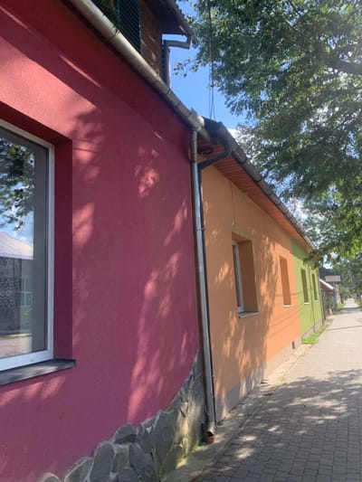 The image captures a sunlit street scene in Yasinia, Ukraine. In the foreground and extending to the left, a building with a reddish-pink stucco exterior features a white-framed window on its left side. The lower section of this building's wall is faced with gray, irregularly shaped stone. A silver-colored metal downspout runs vertically along the right edge of this building, leading into a visible drain pipe. Adjacent to this structure, a second building extends into the midground, its facade segmented into distinct color sections: a light orange-brown, followed by a deeper orange-brown, and then a lime green towards the background. Multiple white-framed windows are visible on this multi-colored building. To the right, a paved sidewalk composed of rectangular gray pavers stretches into the distance, partially shaded by the dense foliage of mature green trees that line the street. The sky above is clear and blue, with sunlight casting distinct shadows of tree branches onto the building walls and sidewalk. No individuals are present in the frame.