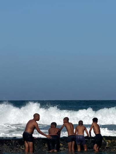 Six shirtless boys, who appear to be dark-skinned, are on a rocky shore at Muizenberg, South Africa. Waves crash against the shore near them. They are positioned along the edge where land meets water. The sky is a clear blue.