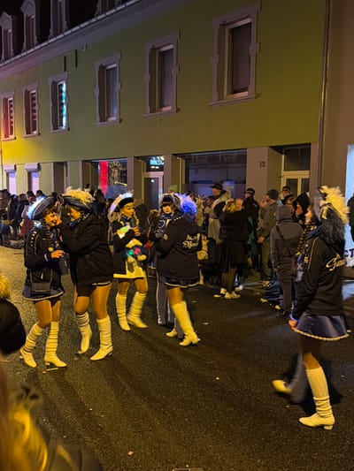 This image depicts a nighttime street scene in Wasserbillig, Luxembourg, featuring a large gathering of people. In the foreground and midground, a group of approximately five to seven females are present, dressed in matching or similar performance costumes. Their attire includes dark navy blue or black jackets, short blue skirts with white trim, opaque tan hosiery, and white knee-high boots or ribbed socks. Several individuals wear distinct headpieces adorned with white or blue faux fur, some of which are illuminated with blue light. One female in the mid-ground holds a small, glowing multi-colored plush toy. Text visible on some jackets includes "KCK" and "Klein Macht" on one, and "K.G. Gem" on another. These individuals are standing on the dark asphalt street, which is scattered with small pieces of white and pink confetti.

In the background, a two-story building with a light olive-green facade features multiple rectangular windows with light-colored frames. Several upper-floor windows are internally lit, one showing a blue light and another a red light. The ground floor comprises storefronts or entrances. One window displays a mannequin dressed in a dark uniform with gold trim. The number "54" is affixed to the wall next to a ground-floor entrance. A dense crowd of people, dressed in various casual winter garments including jackets, hats, and scarves, occupies the background areas of the street and sidewalks. The scene is illuminated by artificial light sources from streetlights and the building's interior. Individuals in the crowd are primarily standing and engaging in informal interactions.
