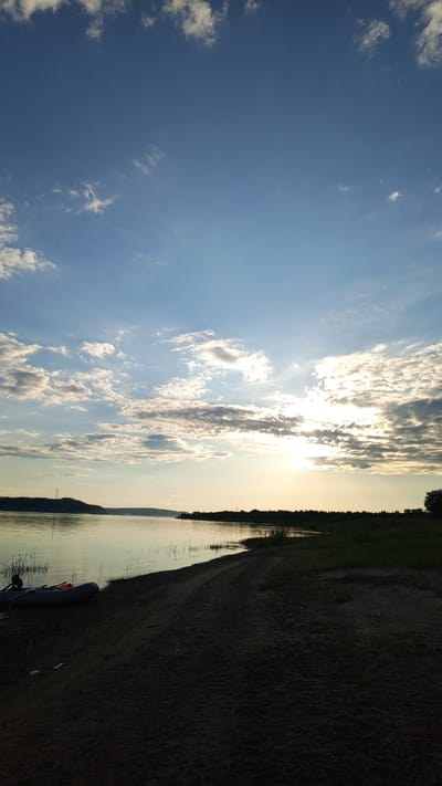 This image captures a wide outdoor landscape scene at either sunrise or sunset over a body of water in Шолья, Russia. The sky occupies the upper two-thirds of the frame, displaying a gradient from deep blue at the zenith to lighter blue and yellow hues near the horizon where the sun is positioned on the right. Numerous white and grey scattered cumulus clouds are visible, particularly concentrated around the sun, which appears as a bright, partially obscured orb casting a radiant glare.
Below the sky, a calm body of water, likely a river or large lake, stretches across the middle ground, reflecting muted tones of the sky. The distant left bank features a dark, undulating landmass with a visible transmission tower or similar utility structure on its summit. The distant right bank is lined with dense, dark green foliage, possibly trees or bushes.
The foreground consists of a sandy or dirt shoreline, appearing dark due to being in shadow relative to the sun's position. A clear, unpaved track or path runs along this sandy bank. On the left side of the foreground, an inflatable boat, grey in color, is beached on the sand. An orange object, possibly a life vest or a piece of equipment, is visible within the boat. Sparse, tall green vegetation grows near the water's edge in the immediate foreground, particularly on the left. No individuals are visible in the scene. Small, dark specks, possibly birds or insects, are discernible in the sky.