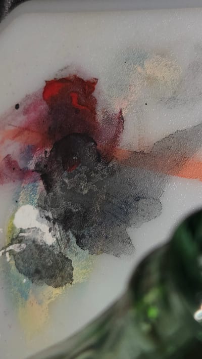 The image shows a close-up of a paint palette with various colors mixed together. Prominent colors include red, black, white, and yellow, with some orange and blue blended in. The palette appears to be made of a light-colored, possibly translucent material. The background is blurry, showing some green foliage, indicating an outdoor or studio setting. The exact location in Lompoc, United States of America is not discernible from the image.