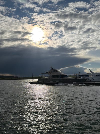 The image depicts a harbor scene in Molodezhnyi, Russia, under a dramatic sky. The sun is partially obscured by dark, heavy clouds, casting a bright, speckled reflection on the choppy water. Several boats are docked along a pier. The largest vessel in the foreground is a yacht with a light-colored hull and a darker lower section. It has multiple windows and a mast. To its right, another white boat with a sail is docked. In the background, a more distant white boat is visible. The shoreline is lined with trees and some low structures. The overall atmosphere is somber due to the stormy clouds, yet illuminated by the sunlight breaking through. No people are visible in the frame.