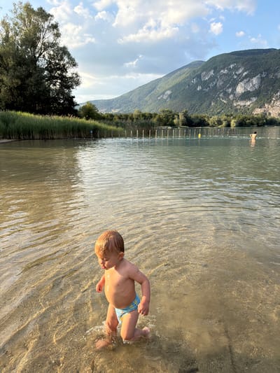 A blond-haired toddler wearing blue swim trunks walks through shallow water near the shore of a lake. The child is facing to the left, with their head turned slightly down and to the right. The water is clear and reflects the sunlight, revealing the sandy bottom. In the background, a person is standing in the water further from the shore, and a line of buoys and poles mark a swimming area. Beyond the lake, tall green reeds line the shore, backed by a dense forest. Majestic green mountains rise in the distance under a partly cloudy sky. The image appears to be taken in the late afternoon, with the golden light illuminating the scene. The location is likely the lake in Novalaise, France, given the presence of mountains and a lake.