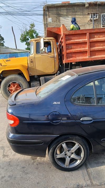 The scene depicts a yellow dump truck with an orange bed parked alongside a dark blue "UNIQUE"-branded car. The truck's cab has a visible driver wearing a blue shirt. The surrounding environment appears to be an urban area in Barcelona, Venezuela, indicated by the presence of buildings, power lines, and paved roads. The objects of significance are the truck, the car, and the urban environment. Actions are limited to the parking of the vehicles and the presence of the driver in the truck.