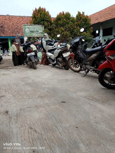 Several people, including children in uniform and women wearing hijabs, stand near parked motorcycles. The motorcycles are lined up on a concrete surface, possibly a parking area. A building with a red tile roof is visible in the background, along with some trees with reddish foliage. The image also has text at the bottom indicating the device used to take the photo.