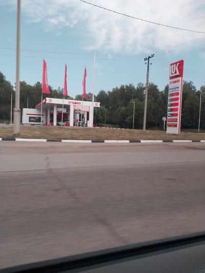The image shows a gas station in Баюновские Ключи, Russia. The gas station has a red and white sign with the logo "ЛУКОЙЛ" and prices for fuel displayed on it. There are several red flags flying in front of the gas station. The scene appears to be outdoors on a clear day with blue skies and some clouds. Trees are visible in the background, suggesting a rural or semi-rural setting. The photograph seems to be taken from a moving vehicle, as indicated by the motion blur on the road in the foreground. The time of day appears to be daytime, likely late morning or afternoon. No people are visible in the image. The overall impression is of a functional, well-lit gas station on a typical day.
