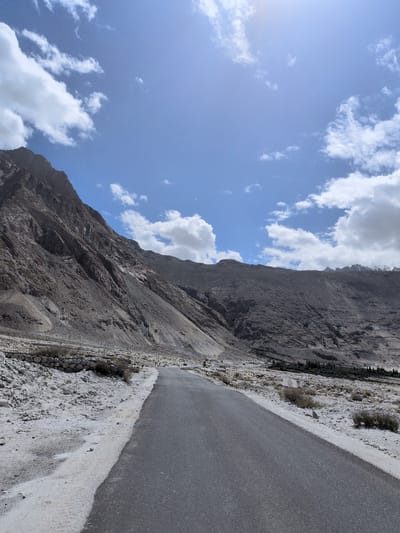 The image depicts a scenic landscape with a paved road leading towards rugged, mountainous terrain. The sky is a vibrant blue with scattered white clouds, and the sun is shining brightly, casting subtle lens flares in the upper portion of the image. The mountains are imposing, with stark, rocky slopes displaying shades of brown and grey. Vegetation is sparse, consisting of low-lying bushes and some scattered trees in the distance, indicative of a dry, arid environment. The ground adjacent to the road is covered in sand and small rocks. There are no people clearly visible in the scene, but there appear to be some faint human figures in the distance on the side of the road. Power lines and poles are visible along the side of the road, suggesting human infrastructure in the otherwise natural landscape. The overall impression is one of a remote and somewhat desolate, yet beautiful, mountainous region.