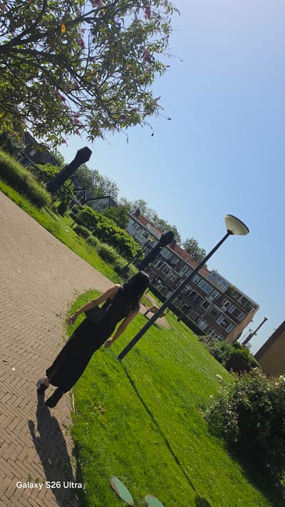 An individual, viewed from behind, walks away from the camera on a brick pathway. They are wearing a floor-length black dress and a black cross-body bag. The person's shadow is cast on the pathway to their left. To the right of the pathway is a lush green lawn. In the background, several apartment buildings with orange-tiled roofs are visible, surrounded by trees and greenery. A lamppost stands near the pathway. The sky is a clear, bright blue. The image appears to be taken on a sunny day in the city of Vlaardingen, Netherlands, given the "Galaxy S26 Ultra" watermark, which is a common device used in that region.