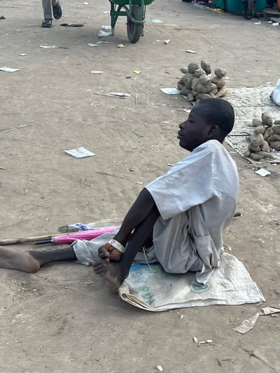 The image depicts a young male sitting on a white sack on the dusty ground of Oko Amakom, Nigeria. He is wearing a light-colored tunic and is positioned with his left leg bent and his right leg extended forward. A pink umbrella lies next to him on the ground, along with a wooden stick. A white wristband with a red heart design is visible on his left wrist. In the background, several piles of tubers are stacked on the ground, and a green wheelbarrow is partially visible. The environment appears to be an outdoor market or thoroughfare with scattered debris on the ground.