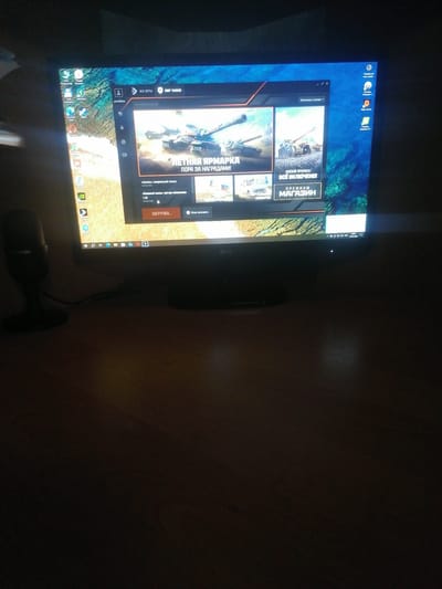 This is a low-light, angled photograph of a computer monitor displaying the "World of Tanks" game launcher. The monitor is situated on a dark surface, likely a desk, in a dimly lit room. The screen shows various game-related content, including images of tanks, a "Summer Season" banner, and options to download or launch the game. To the left of the monitor, a black microphone with a silver brand name "Trender" is visible. The background of the computer desktop is a scenic image. The overall scene is dark, with the monitor being the primary source of light. No people are visible in the image. There is no specific information or context provided within the image itself that would indicate its location in Yelsk, Belarus.