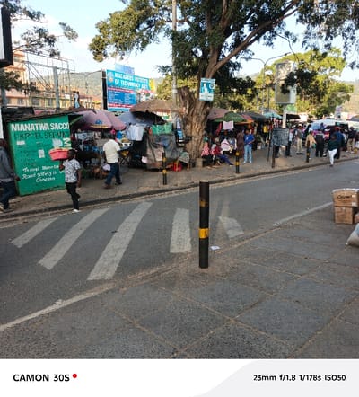 This media file captures a vibrant street scene in Machakos Central ward, Kenya, likely depicting a market area or a busy thoroughfare. The setting appears to be an urban or semi-urban environment under bright daylight, suggested by the clear sky and even illumination.
The main subjects include numerous people of various ages, both walking and congregating. Some individuals are seated on low stools near market stalls, while others are standing or moving along the pavement and street. The scene is bustling, implying activity such as commerce, commuting, or social interaction.
Prominent objects include several informal market stalls with umbrellas providing shade, displaying various goods, although specific items are not clearly discernible. A green kiosk or structure on the left features text. There are mature trees with dense foliage, providing shade to parts of the street. A zebra crossing is visible on the asphalt road, and black and yellow bollards delineate areas of the street. Cardboard boxes are stacked on the right side of the road. In the background, buildings and distant hills are visible.
Visible text includes:
* On the green structure: "MANATWA SACCO PARCEL SERVICE" and partially "Travel Manatw Road safety is Concern."
* On a blue sign mounted on a pole: "MACHAKOS INSTITUTE OF TECHNOLOGY MIT."
* In the bottom left corner: "CAMON 30S," indicating the camera used.
* In the bottom right corner: "23mm f/1.8 1/178s ISO50," which are the EXIF data of the photograph.
The overall impression is one of a busy, lively public space, characteristic of daily life in a Kenyan town or city.