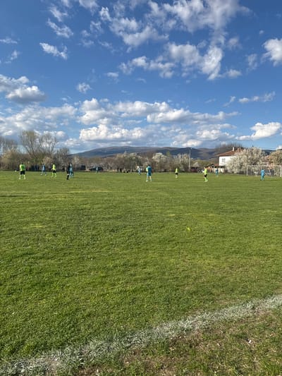 The image depicts a soccer field in Zhabokrat, Bulgaria, with several players in yellow and blue uniforms practicing. The field is green with a white sideline visible on the right. A soccer goal is positioned on the far end of the field. In the background, there are trees, a building, and a mountain range. The sky is blue with white clouds.