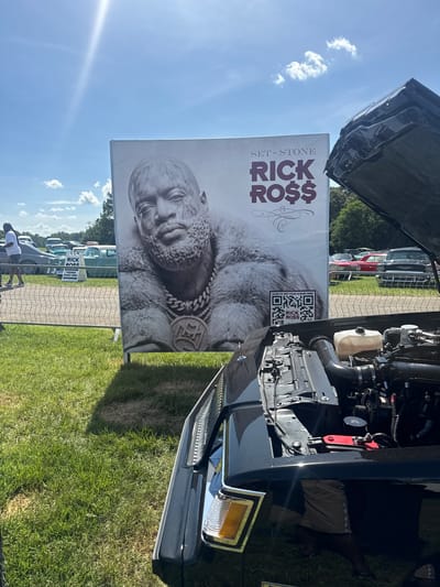 In Fairburn, Georgia, an outdoor event, possibly a car show, is underway on a sunny day. In the foreground, the open hood of a black car reveals its engine components. To the left, a large billboard displays a black and white portrait of rapper Rick Ross, promoting his album "Set 'n Stone" and featuring a QR code for album access. Behind the billboard, several vintage cars are parked on a grassy field. A fence separates the viewing area from the parked vehicles. People are visible in the background, some walking and others near the cars. The sky is clear blue with a few scattered clouds.