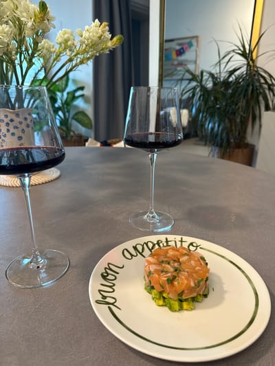 Two wine glasses are on a table, one in the foreground and one further back. Each glass is filled with red wine. In the background, a plate holds a salmon tartare dish. The plate has green writing on it that says "Buon appetito". To the left of the wine glasses, a vase holds white flowers. The table appears to be a dark grey surface, possibly a dining table. The scene is indoors, with a blurred background suggesting a home setting. The lighting appears to be natural, perhaps from a window. There is no indication of time of day, weather, or specific emotions. The location is identified as منجا, Jordan.