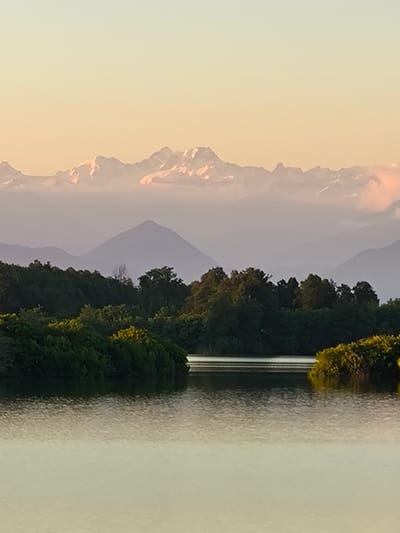 This is a vertical, eye-level, landscape photograph capturing a serene natural scene. The image is dominated by layers of mountains fading into the distance under a soft, pale orange sky, suggesting either sunrise or sunset. The uppermost mountains are snow-capped and appear rugged, kissed by the warm light of the setting or rising sun. Below them, successive ranges of mountains are visible, becoming progressively hazier and bluer with distance, creating a sense of depth and scale.
In the foreground, a calm body of water reflects the soft sky and the silhouettes of trees. The water's surface is smooth, with subtle ripples indicating a gentle breeze. Lush green trees line the banks of the water, forming a dense treeline. Some of these trees have yellow-green foliage, possibly indicating autumn colors or sunlight catching them. The overall mood is peaceful and tranquil, evoking a sense of natural beauty and quietude. There are no people or man-made objects visible in the frame, emphasizing the untouched wilderness of the setting. The time of day is clearly indicated by the warm, diffused light and the long shadows or lack thereof. The weather appears clear, with soft light bathing the landscape. The location context provided (Baghlan, Abkhazia) suggests a mountainous region with water bodies. No text is visible in the image.