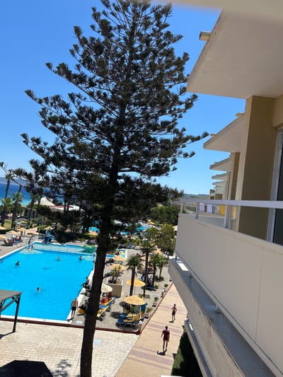 This is an outdoor, high-angle, full shot of a resort with a large, bright blue swimming pool in the foreground, and the Aegean Sea in the background. The resort features palm trees, lounge chairs with yellow umbrellas, and people swimming or relaxing. A tall, dark-barked pine tree with sparse foliage is in the center of the frame, partially obscuring the view of the resort. The building on the right is a multi-story hotel with balconies. The sky is clear and blue. The image appears to be taken during the daytime.
