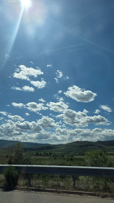The image depicts a scene outdoors, likely captured from a moving vehicle on a road. The sky is a vibrant blue, dotted with numerous puffy, white cumulus clouds. In the lower-left corner, the sun shines brightly, creating a lens flare effect. The lower portion of the image shows a guardrail along the roadside, with vegetation and a distant view of a hillside or low mountain range. The location cannot be determined to be Mursalevo, Bulgaria from this image.