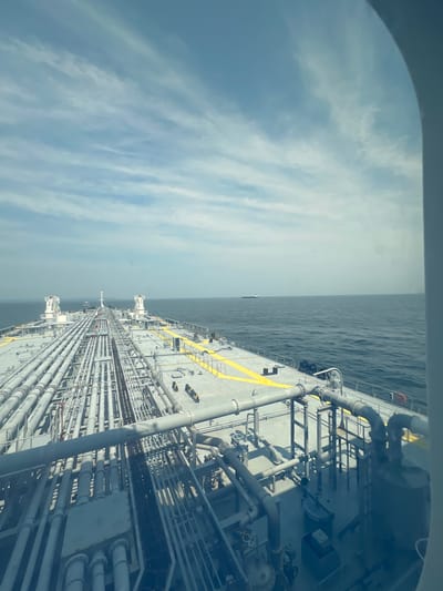 This media file shows a view from a window looking out over the deck of a large cargo ship. The ship's deck is covered in a complex network of pipes and conduits, painted in various shades of gray with yellow safety markings. The ship appears to be at sea, with the vast expanse of the ocean stretching to the horizon. In the distance, another ship is visible. The sky is partly cloudy, with streaks of white clouds against a pale blue background, suggesting daytime. The lighting indicates it's likely late morning or early afternoon. The scene conveys a sense of industrial activity and vastness, with the powerful presence of the ship against the backdrop of the sea and sky. There is no discernible activity of people in the image, but the infrastructure suggests an ongoing operation, likely related to the transport of goods. The location is identified as Saint John, Canada, implying the ship may be docked or en route to or from this port.