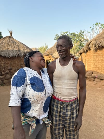 An older man with a tank top and plaid pants smiles at a woman wearing a patterned white and blue shirt. The woman, with a black headband, also smiles broadly. Behind them, a traditional thatched-roof hut is visible in a rural setting. The sky is clear. Both individuals appear to be sharing a joyful moment together.