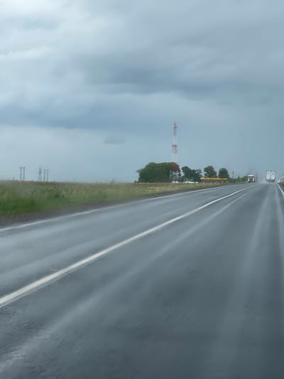 The image depicts a road, likely a highway, located in Кокино, Russia, under overcast and possibly rainy conditions. The asphalt road surface appears wet and reflective, with a clear white line marking the lane division. On the left side of the road, there is a grassy shoulder followed by a landscape containing a communication tower with red and white stripes, some trees, and power lines in the distance. The sky is heavily clouded, suggesting poor weather. There are a few vehicles visible on the road ahead in the distance.