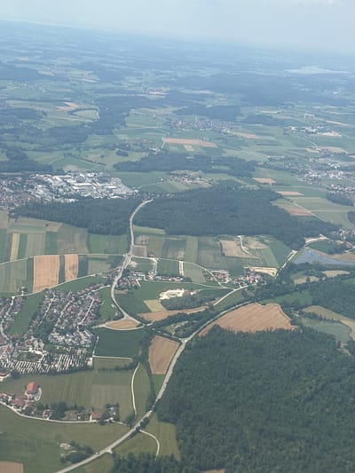 This is an aerial photograph of the landscape surrounding the city of Muntigl, Austria. The image showcases a rural setting with a patchwork of agricultural fields, forests, and a small urban area. The fields exhibit varied crop types and colors, indicating different stages of growth or cultivation. Several roads wind through the landscape, connecting scattered houses and buildings. A prominent feature is a large industrial complex visible in the upper left portion of the image, characterized by numerous buildings and parking areas. The terrain appears gently rolling, with trees and vegetation covering significant portions of the area. The sky is overcast with a hazy, diffused light, suggesting a cloudy day. No people or animals are discernible in the image. The overall impression is one of a developed rural area with agricultural and industrial activity.