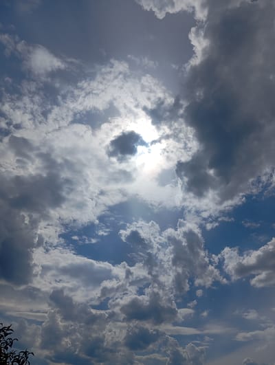 The image shows a dynamic sky filled with a mix of dark, dramatic clouds and brighter, wispy ones, with the sun peeking through. The sunlight creates a halo effect and highlights the textures of the clouds. Some rays of light are visible, suggesting a partly cloudy day. The blue sky is visible in patches between the clouds. In the bottom left corner, there are dark silhouettes of tree branches, possibly indicating an outdoor setting. The overall impression is one of a vibrant and somewhat dramatic sky, typical of a day with changing weather. The time of day appears to be daytime, likely late morning or afternoon, given the brightness of the sun. There is no visible text in the image. The location context provided is Buzke, Ukraine.