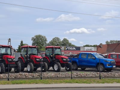 An outdoor, daytime, eye-level medium shot captures a row of tractors and two pickup trucks parked in a lot in Ślesin, Poland. The foreground features a gray asphalt sidewalk with a thin strip of green grass separating it from a chain-link fence. Beyond the fence, a gravel-covered area displays several red tractors and a blue pickup truck. Behind the vehicles, a building with a white facade and a red-tiled roof is visible, with a black and white sign that reads "STIGA SKLEP TECHNIC DAM-POL." The sky above is a light blue with scattered white clouds. There are no people visible in the scene.