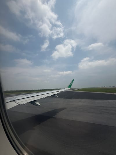 This image captures a view from an airplane passenger window, oriented vertically, likely taken during ground operations at an airport. The aircraft's white wing, featuring a distinct green wingtip with the text "VIVA" in white capital letters, extends diagonally across the middle of the frame. Portions of the airplane's engine nacelles are visible beneath the wing.
To the left of the wing, the sky is partly cloudy, displaying patches of light blue interspersed with white cumulus clouds. The natural lighting suggests it is daytime, possibly late morning or early afternoon, with bright but not harsh illumination.
Below and to the right of the wing, a dark asphalt runway is visible, showing some lighter markings. This paved surface is bordered by a vibrant green grassy area, suggesting the aircraft is either taxiing, awaiting takeoff, or has just landed. The curved frame of the airplane window is partially visible in the bottom right corner of the image.
There are no people visible in the scene. Given the location context of Apodaca, Mexico, the airport is likely Monterrey International Airport (MMTY), which serves as a hub for Viva Aerobus. The image conveys a sense of travel and the commencement or conclusion of a flight.