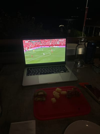 An outdoor patio at night in Gersau, Switzerland. The scene is dominated by a laptop displaying a soccer match. The laptop screen shows the score "33:32 SUI 0 | 0 CAN". A red plastic cutting board with diced cheese and cubed meat is in the foreground. A glass bottle and a can of beverage are on the table next to the laptop. Trees are visible in the background.