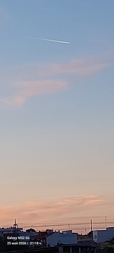 The image shows a blue sky with wisps of pink clouds near the horizon. A contrail left by an airplane is visible in the upper-left quadrant of the frame. Along the bottom-right, there are buildings and other structures in an urban or suburban setting. The photo seems to be taken with a Samsung Galaxy M52 5G on May 25, 2026, at 21:18. The information indicating the location in l'Alcúdia de Crespins, Spain, is not directly discernible from the image.