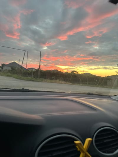 The image shows a reflection of a dramatic sunset on a car windshield. The sky is filled with vibrant clouds in shades of orange, pink, and gray, illuminated by the setting sun. The sun itself is a bright yellow glow on the horizon, casting a warm light over the landscape.
The reflection on the windshield reveals a roadside scene. A road stretches into the distance, bordered by vegetation and some buildings. There are also utility poles visible. The interior of the car is visible on the right side of the image, with a dashboard featuring air vents and a yellow hanging ornament.
The time of day is clearly sunset, given the colors in the sky and the warm lighting. The weather appears clear, allowing for a spectacular sunset display. The location context of Miatli, Russia, suggests a rural or semi-rural setting. There is no visible text in the image. The overall mood conveyed by the image is one of natural beauty and peacefulness.