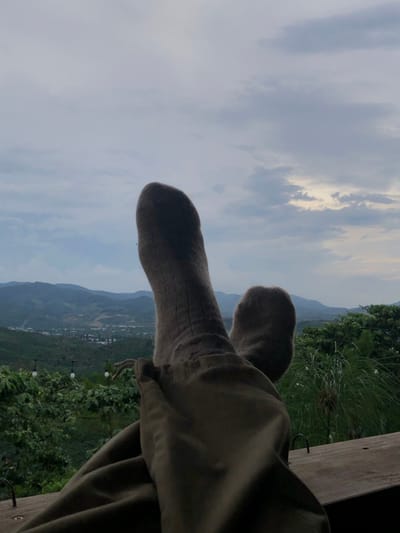 The image features a person's feet, clad in thick, grey socks, resting on a wooden surface. The feet are positioned in the foreground, with one foot extending forward and the other tucked in slightly. The socks have a ribbed texture. The person is wearing olive-green trousers, the fabric of which is draped over the legs.
The background reveals a scenic view of a valley, likely in Thôn 4, Vietnam, with rolling hills covered in lush green vegetation. A small village or settlement is visible nestled within the valley. The sky above is partly cloudy, with soft, diffused light suggesting either early morning or late afternoon. The overall atmosphere is calm and serene. There are no visible signs of text in the image. The focus is on the relaxed posture of the person and the natural beauty of the surroundings.