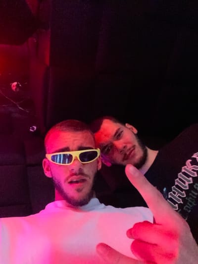 Two males are present in a dimly lit, indoor environment with atmospheric red and pink lighting, likely a nightclub or lounge in Пампорово, Bulgaria. The subject in the foreground is a light-skinned male with a short buzz cut and stubble, wearing a white t-shirt and narrow, reflective yellow sunglasses. He is looking directly at the camera, with his left hand raised in a "rock on" gesture. Behind and to his right, a second light-skinned male with dark hair and a beard is partially visible, wearing a dark t-shirt with indistinct light-colored text. He is also looking towards the camera. The background consists of dark, possibly upholstered walls, with a dark, reflective surface visible on the left side, showing a faint red reflection and a small circular metallic object.