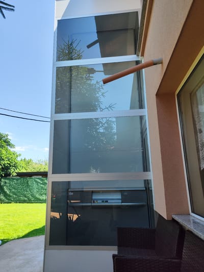The image displays an outdoor residential scene in Brestnik, Bulgaria. A multi-story external elevator shaft, constructed with large rectangular glass panels set within a light-colored metal frame, occupies the left side of the frame. The glass surfaces reflect sections of a nearby evergreen tree and the clear blue sky. The lowest visible section of the elevator's interior features a control panel with a green '0' button, a green '1' button, and a red stop button, situated above a horizontal metallic bar. The interior behind the panel appears to be a dark grey material.

To the right, a section of an adjacent house is visible, presenting a textured, light peach-colored stucco facade. A window with a white frame and a white lace curtain is present on the house wall, positioned above a granite-patterned windowsill. A brown PVC pipe, fitted with a grey elbow joint, extends horizontally from the house wall, casting a shadow.

In the lower right foreground, dark brown woven outdoor furniture, consistent with chairs or a sofa, is partially visible. The area to the left of the elevator shaft comprises a manicured green lawn, beyond which a green mesh or hedge fence borders dense green deciduous trees. The sky above is bright blue with scattered white clouds, traversed by two dark electrical wires in the upper left quadrant. No human subjects are present in the scene.