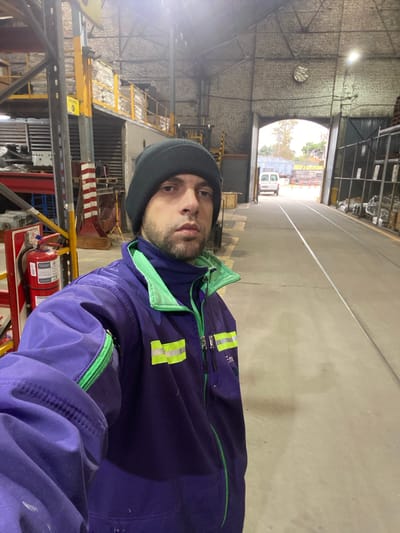 A man wearing a grey beanie and a blue and green work jacket with reflective strips is taking a selfie inside what appears to be a large industrial building or warehouse. Behind him, there is a large arched doorway leading to an outside area where a white van is parked. The building's interior has exposed brick walls, a high ceiling with visible structural elements, and what seems to be shelving and equipment. A fire extinguisher is also visible in the foreground. The specific location in Sáenz Peña, Argentina, cannot be determined from the image.