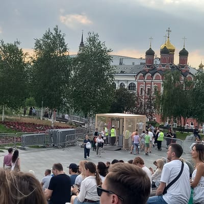 This image depicts an outdoor gathering in a public space, likely a park or plaza, with a church building in the background. The church features multiple domes, with one prominent golden dome and several smaller, darker domes. The building is ornate, with red walls and white decorative elements. The sky is overcast with hints of warmer light peeking through.
In the foreground and midground, numerous people are present, some standing and others seated. Many individuals appear to be spectators, facing towards an unseen point of interest or event. Several individuals in bright yellow vests are visible, suggesting they may be event staff or security. There are also trees with dense foliage scattered throughout the scene, and metal barriers that might delineate areas or pathways. A wooden structure with a slatted design is also present, possibly a kiosk or a stage element. The ground is paved, and there are flowerbeds with red flowers in the middle ground. The overall atmosphere suggests a public event or festival.