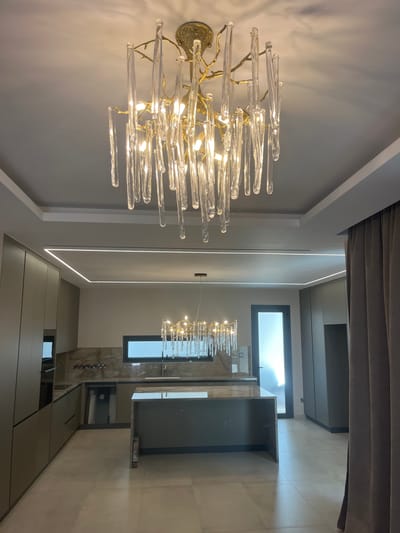 This is a full-shot, eye-level, interior photograph of a modern kitchen in Moni Community, Cyprus. The kitchen features a large island with a grey countertop, custom cabinetry in a muted olive green, and a sleek marble backsplash. Two prominent chandeliers, one hanging above the island and another suspended from the ceiling, illuminate the space with a warm glow. The room also includes recessed ceiling lights and a large window offering a view of the outdoors. A tall, dark-framed door is visible in the background, leading to an area with natural light. The floor is tiled in a neutral tone. The photograph is well-lit and captures the contemporary design elements of the kitchen.